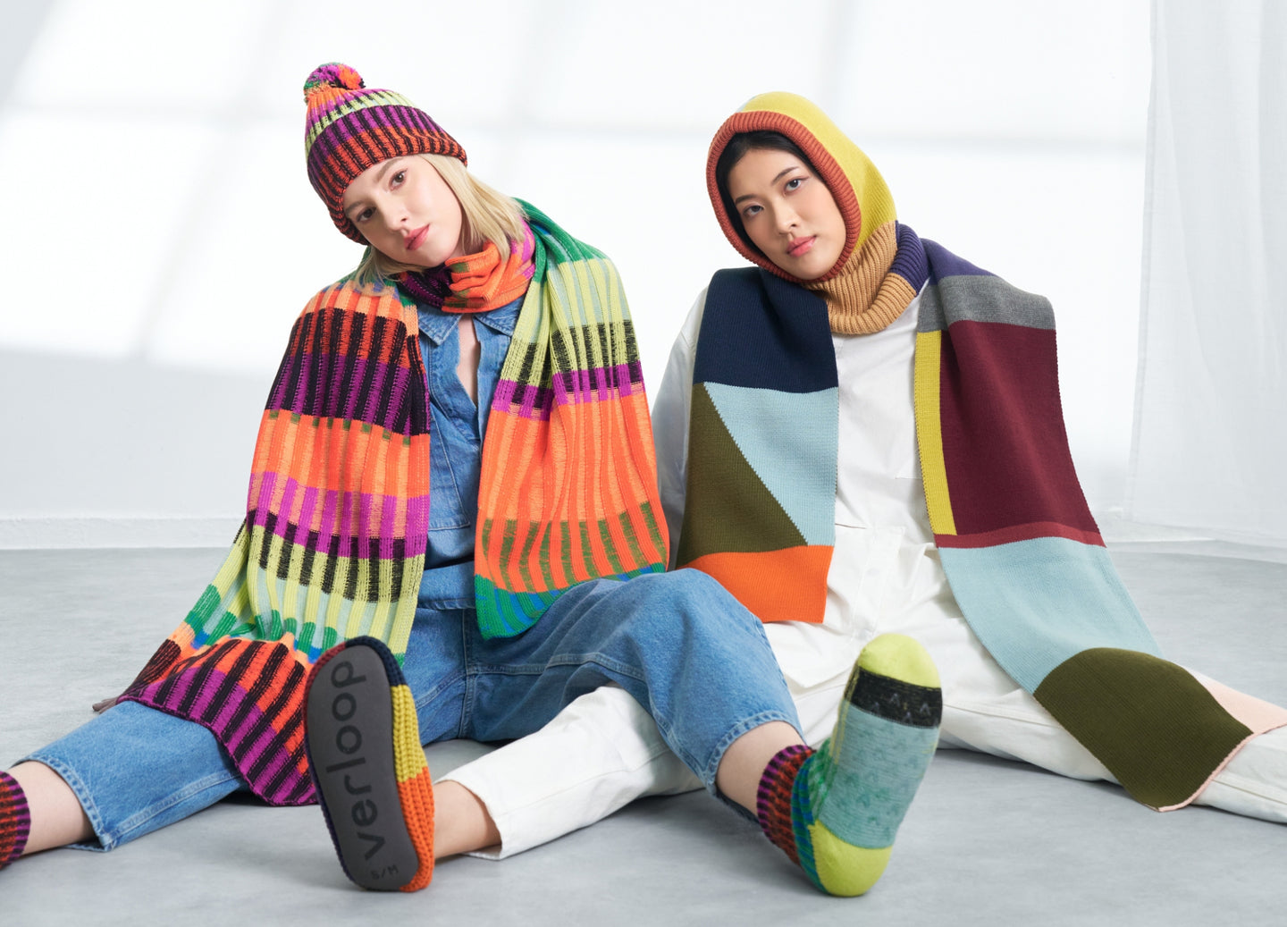 two models wearing matching sets of oversized graphic knit scarves with coordinating beanies and hoods 